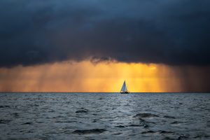 Lonely sailboat sailing in the ocean at the approaching storm and raining clouds at sunrise, English channel, near French shores